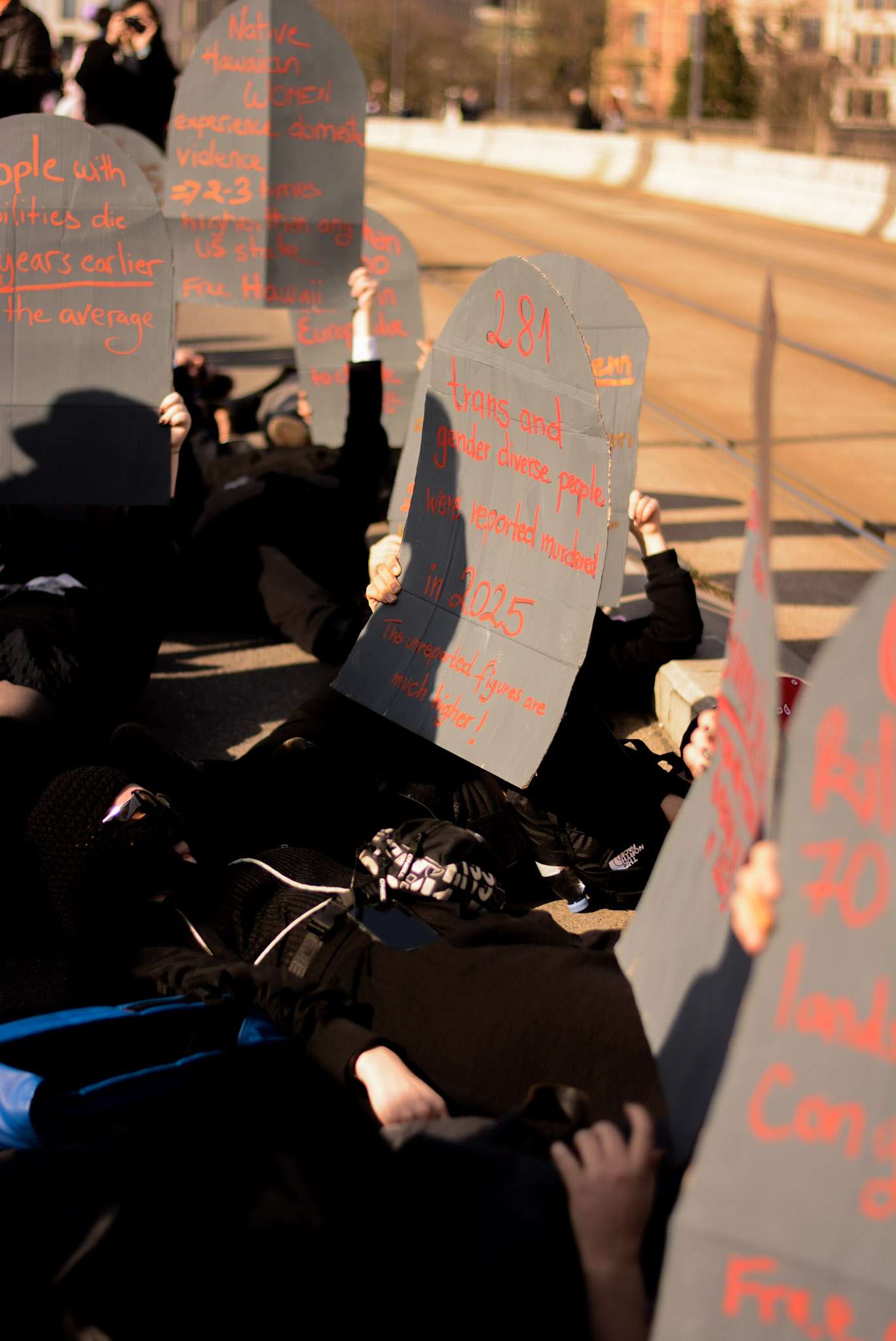 Picture of people lying on the ground holding up cardboard gravestones with statistics on violence against marginalised people.